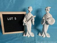 Both white Geisha sculptures positioned on a blue cloth backdrop next to a LOT 5 sign.