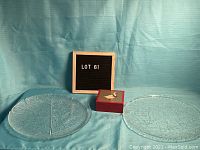 Photo showing two glass serving plates and a boxed coaster set against a teal backdrop, together with a sign labeled LOT 61