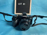 Front view of the Minolta Maxxum 3000i 35mm film camera with attached lens and neck strap, set against a blue background with a black letter board showing 'LOT 81'.