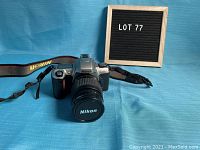 Front view of Nikon N65 camera with Nikkor lens and Nikon strap against blue background.