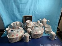 Full dish set arranged on blue surface with light blue background, showing all components including casserole dishes, plates, cups, teapot, serving dishes, salt and pepper shakers