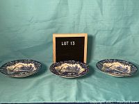 Three matching English Ironstone plates in blue and white with detailed rural scenes, laid out against a blue cloth backdrop with a wooden 'LOT 13' sign behind them.
