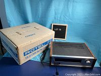 Photo showing the ROTEL RT-320 AM/FM Stereo Tuner alongside original cardboard box. The box has fragile and brand markings. The tuner is placed on a blue surface against a blue backdrop.
