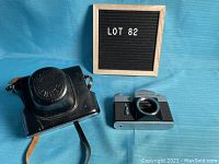 Photo showing the Zenit-B camera body and its black leather carrying case side by side on blue fabric background with a sign reading LOT 82.