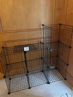 Two black wire shelving units assembled from modular cube grids on a wooden floor against a wooden-paneled wall.