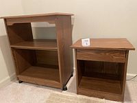 Front view of two wooden side tables against a wall, one larger with two shelves and casters, one smaller with drawer and shelf.