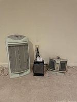 Three Holmes brand portable heaters placed side by side on carpeted floor, showing front and power cords.