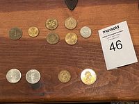 The first photo shows 11 assorted foreign coins laid out on a wooden surface, displaying mixed sizes and colors including silver, bronze, and gold tones.