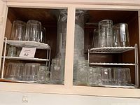 Photo of shelves with over 20 assorted clear glass drinking glasses and mugs arranged on white metal racks beneath a cabinet.