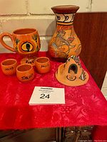 Photo showing six Aztec style ceramic items on red cloth: a pitcher, a mug, three small shot glasses labeled 'SALUD', and a triangular candle holder.
