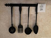 Photo of four black wrought-iron decorative kitchen utensils hanging on a matching wall mount rack, arranged from slotted spoon, fork, ladle, to solid spoon from left to right.