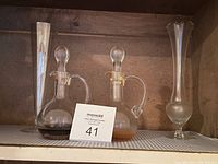 Two glass oil and vinegar cruets with stoppers placed between two tall clear glass vases on a shelf.