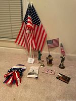 Photo showing three American flags with stands and various small patriotic figurines and flat collectibles arranged on carpeted floor against a wall.