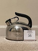 Stainless steel Paul Revere kettle with black handle and spout cover, shown standing on a floor against a beige wall with auction number card 35.
