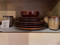 Stack of brown glazed 10.5 inch dinner plates with salad bowl on top and 3 coffee mugs arranged beside stack.