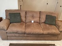 Front view of tan micro-suede couch with three back cushions and two green square pillows, showing soft padded armrests and camel tan color fabric.