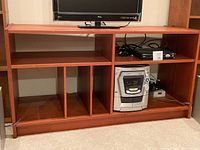 Wooden TV stand with a flat top surface holding a TV and multiple open shelving compartments containing various electronic items (not included in the lot).