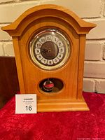Front view of wooden mantel clock on red fabric with a small pendulum visible and white brick background