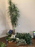 Wide view of assorted artificial plants grouped together on a wood floor against a wall, including a tall tree, leafy bushes, and flowers in pots, vases, buckets, and baskets.
