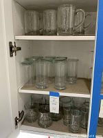 Five clear glass mugs with handles on top shelf, seven small clear glasses on middle shelf, eleven larger glasses on bottom shelf inside white cabinet.