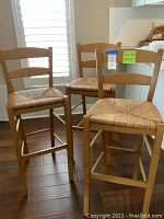 Three wooden bar stools gathered together on wood flooring with white plantation shutters in the background, showing their construction and rush seats.