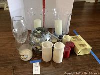 Photo showing assorted candles and vases arranged on a floor including two large clear hurricanes with white candles, spherical mosaic glass vase, battery-operated candles, pillar wax candles in white and red, and a jar candle.