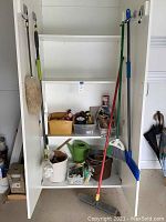 Overall view of white cabinet with two doors, shelves holding multiple cleaning and household items including brooms, buckets, watering can and containers of smaller items.
