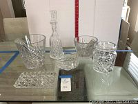 Full view of all six crystal barware and serveware items on glass table showing decanter, ice buckets, bowl, covered jar, and dish arranged together.