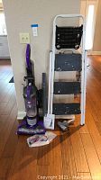 Full view of purple Dirt Devil upright vacuum with hose attachments and a 54-inch white metal step ladder with 3 steps, placed against a wall on hardwood floor.