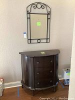 Foyer cabinet with three drawers and decorative hardware beneath a rectangular mirror with a black metal frame and scrollwork.