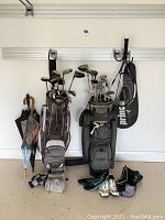 Wide shot showing two golf bags filled with clubs, Prince tennis racket with cover hanging alongside, umbrellas on left, golf shoes and gloves on floor beneath bags.