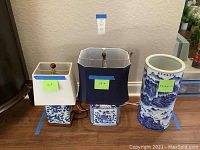 Three items against wall: two porcelain lamps with rectangular lampshades (white and blue) featuring blue dragon and floral motifs, and a cylindrical blue and white porcelain umbrella stand with landscape design