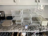 Front view of six white metal wire sliding baskets stacked in two levels with rectangular white plastic basket and turntable on granite countertop under white tiled backsplash