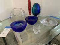 Wide view of all seven glass pieces on a glass table, showing various shapes and colors.