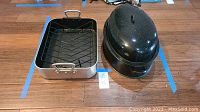 Two roasting pans side by side, one with a removable rack inside and the other with a domed lid on top, placed on a wooden floor.