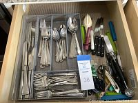 Drawer with stainless steel flatware organized in tray compartments, including knives, forks, spoons, salad forks, and additional kitchen utensils on the right side such as steak knives with black handles and colorful plastic kitchen tools.