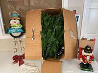 Unassembled artificial evergreen Christmas tree packed in an open cardboard box, metal Christmas penguin yard decoration, and red plaid bow on table.