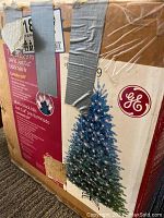 Original cardboard box with manufacturer labels and an image of the decorated tree on front. Labels indicate it's a 7.5 ft pre-lit Just Cut Black Hills Fir with Constant On lighting.