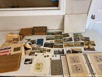Wide view of table displaying multiple vintage postcards, newspaper clippings, old photos, coins, and two wooden cigar boxes.