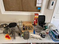 Full view of assorted kitchen items including cookware, flatware, mugs, hand mixer, electric carver, and measuring cups arranged on a counter.