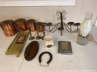 Photo showing five copper nesting canisters, metal wall candle holder with four arms, three brass candlesticks, electric oil lamp-style lamp, white ceramic warmer, horseshoe, framed photo, brass plaque, and other small household items arranged on a surface.