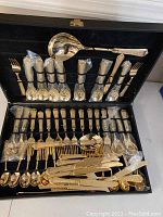Full view of the open case containing the gold-tone flatware service neatly arranged with elastic holders and some pieces wrapped in protective plastic.