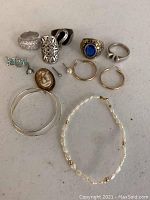 Photo showing 13 jewelry items on a flat pale surface including various rings, stud earrings, hoop earrings, a cameo brooch or pendant, and two bracelets.