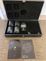 Opened black presentation case displaying four etched lowball whisky glasses and one etched snifter glass, along with a Johnnie Walker branded booklet and aroma wheel card on the floor in front of the case.
