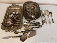 Photo showing two silver-plated serving trays and multiple silver-plated spoons and serving tools with tarnish and wear.