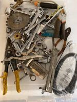 Assorted metal hand tools with yellow-handled locking pliers, circular saw blade, socket wrenches, and various ratchets piled in a box.