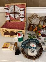 Wicker picnic basket with checkered red and white interior and straps securing white plates and utensils; holiday wreaths and owl figures placed beside it, along with a small snowman 'Happy Holidays' wreath decoration and assorted glass globes with one globe broken.