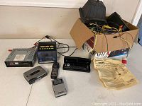Overview photo of vintage electronics and lighting system parts laid out on table, including clock radio, stereo head unit, remote key, cassette player, box with wires, and instruction sheet.