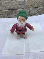Front view of Bashful the Christmas dwarf figurine wearing a red striped shirt, green hat, brown pants, white beard, standing on a white surface.