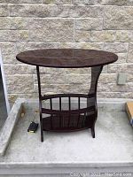Oval dark wood end table with built-in book carrier and oval top surface, photographed from front view outdoors.